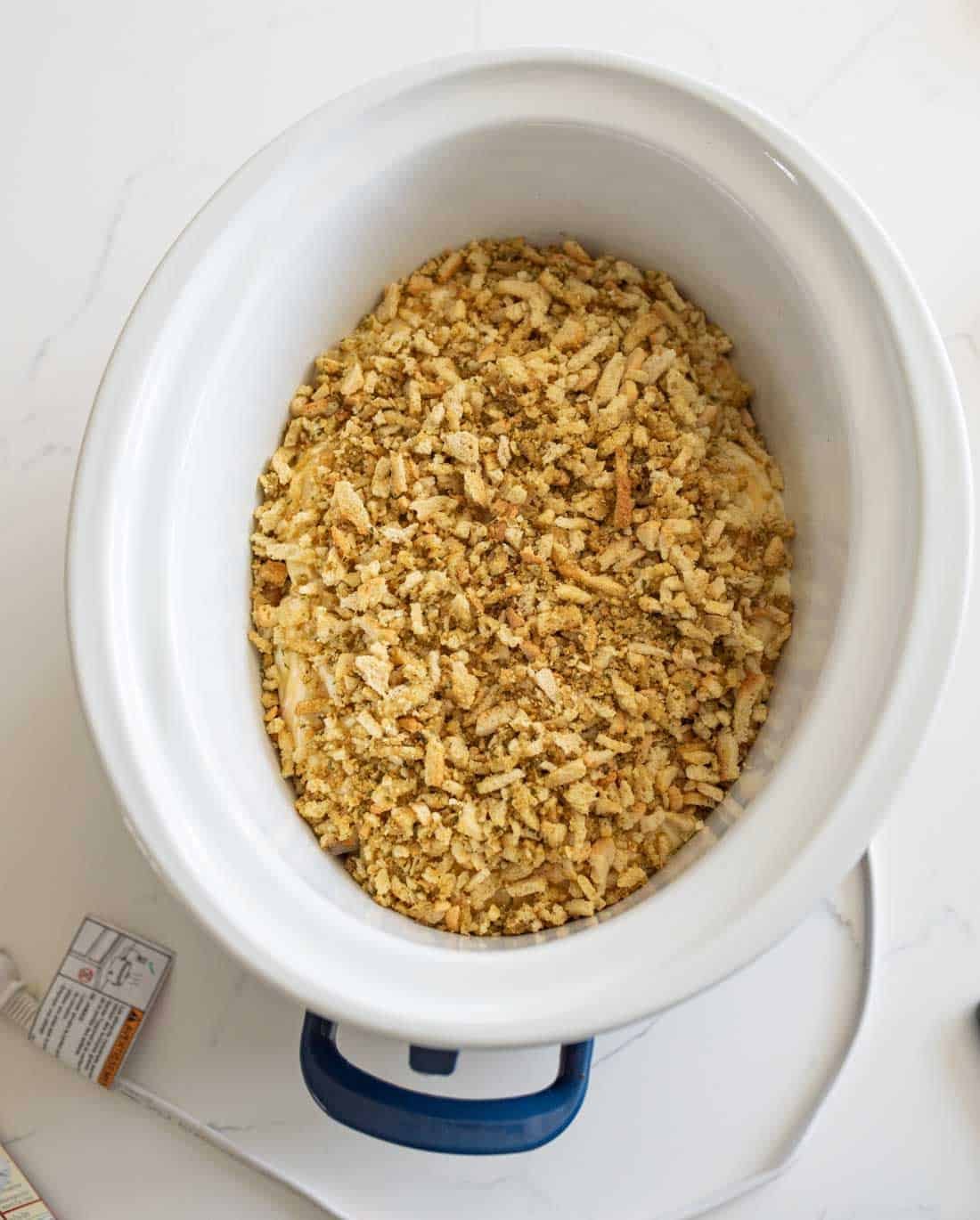 A white slow cooker filled with a crumbly mixture of stuffing or bread crumbs sits on a white countertop, next to a power cord and part of a food package.