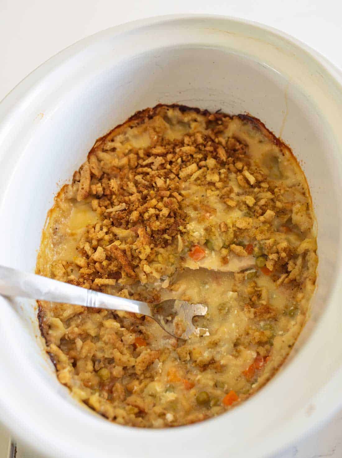 A slow cooker filled with a partially served casserole containing rice, peas, carrots, and a crispy topping, with a metal serving spoon resting inside.