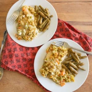 Two white plates with chicken casserole, mixed vegetables, and cooked green beans on a wooden table, with a red patterned cloth underneath. Each plate has a fork.