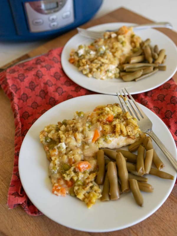 Two white plates hold servings of slow cooker chicken and stuffing casserole with vegetables and a side of green beans. The plates are on a red napkin and a wooden surface, with a blue slow cooker in the background.