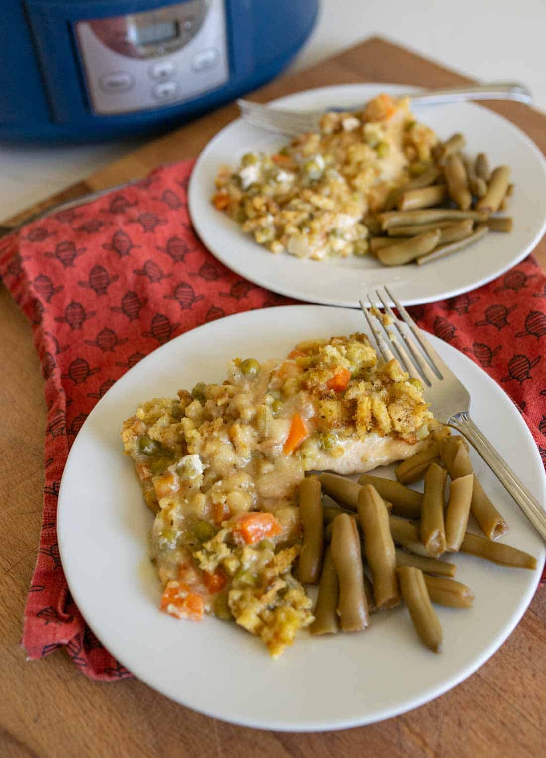Two white plates hold servings of slow cooker chicken and stuffing casserole with vegetables and a side of green beans. The plates are on a red napkin and a wooden surface, with a blue slow cooker in the background.