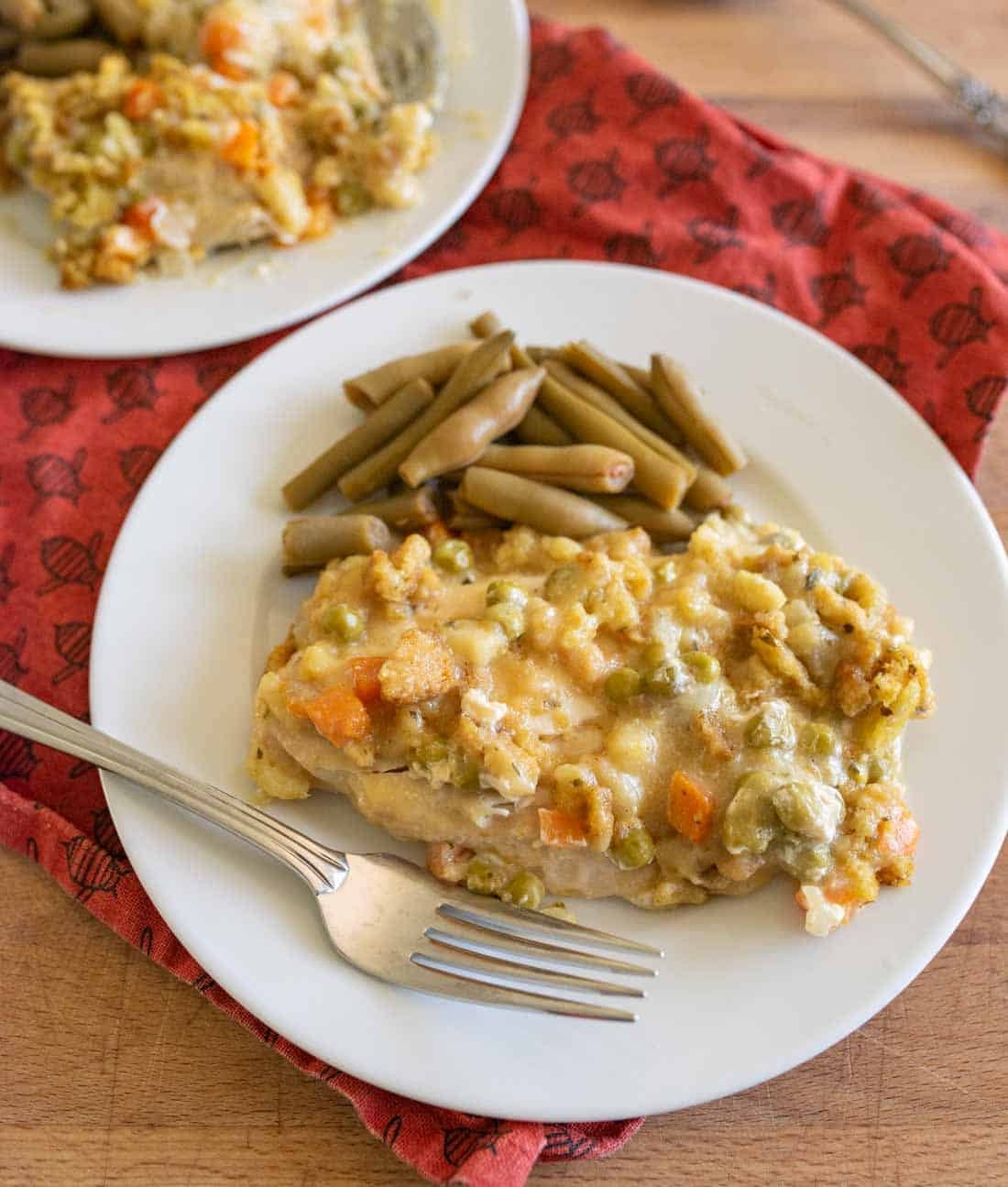 A white plate with a serving of creamy casserole containing vegetables and topped with melted cheese, alongside green beans. A fork rests on the plate, and another plate is partially visible in the background on a red cloth.