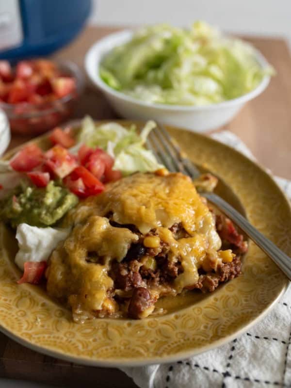 A plate of cheesy taco casserole with ground beef, beans, and corn, served with shredded lettuce, diced tomatoes, guacamole, and sour cream, on a yellow patterned plate with a fork.