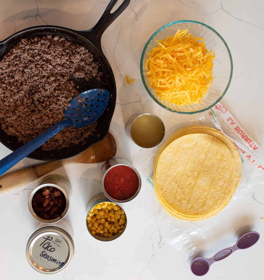 A skillet of cooked ground beef, shredded cheddar cheese, corn tortillas, taco seasoning, canned beans, corn, salsa, and a small bowl of broth, all arranged on a white countertop.