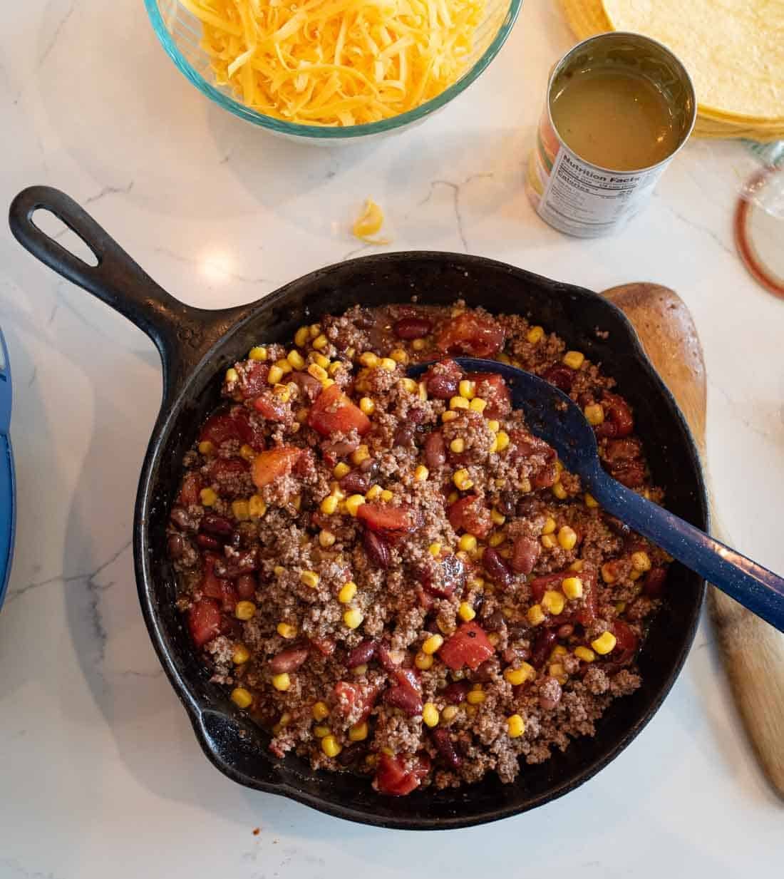 A cast iron skillet filled with cooked ground beef, corn, tomatoes, and beans sits on a countertop next to a bowl of shredded cheddar cheese, an open can of green chilies, and a wooden spoon.