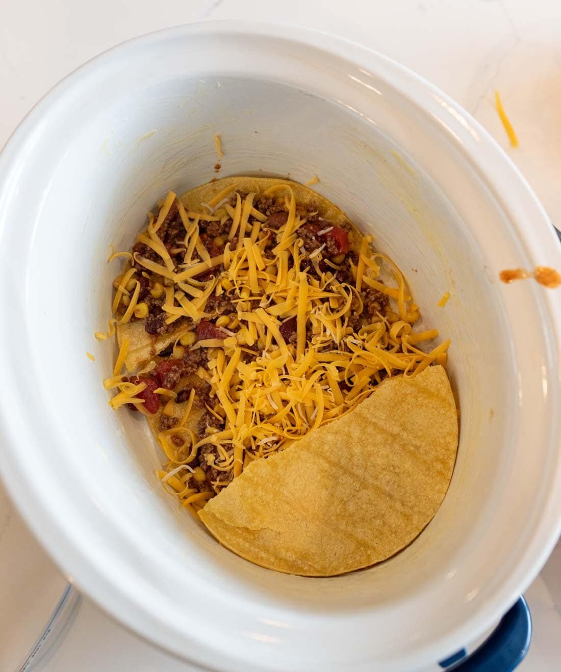 A slow cooker with a layer of ground beef, beans, and shredded cheese topped with corn tortillas, assembling a taco casserole.