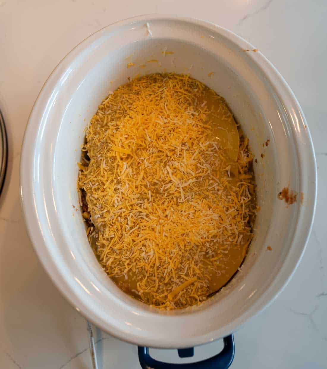 A slow cooker filled with a casserole topped with a layer of shredded cheese, sitting on a white countertop.