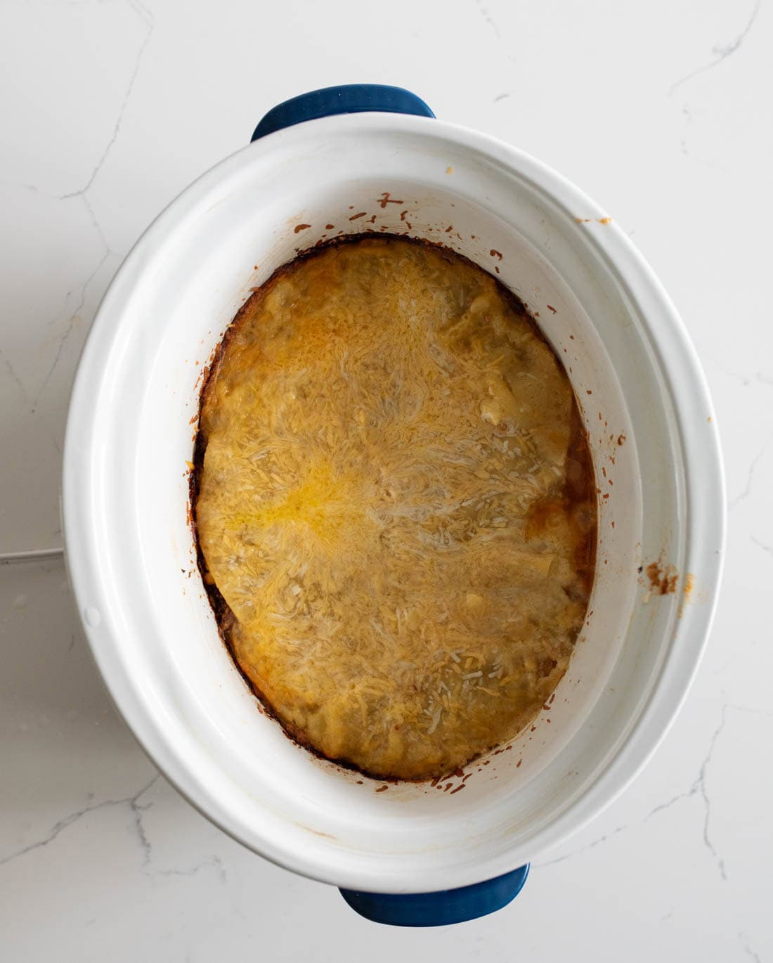 A white oval slow cooker filled with a baked dish topped with melted cheese, seen from above, set on a light marble surface.