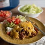 A plate with cheesy slow cooker beef enchiladas, corn, melted cheese, and sides of chopped tomatoes, guacamole, sour cream, and lettuce. A bowl of shredded lettuce and another with diced tomatoes are in the background.
