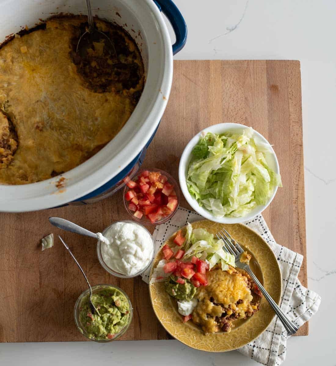 A serving of cheesy casserole on a yellow plate with guacamole, sour cream, lettuce, and diced tomatoes. Nearby are bowls of toppings and a casserole dish with remaining food on a wooden board.