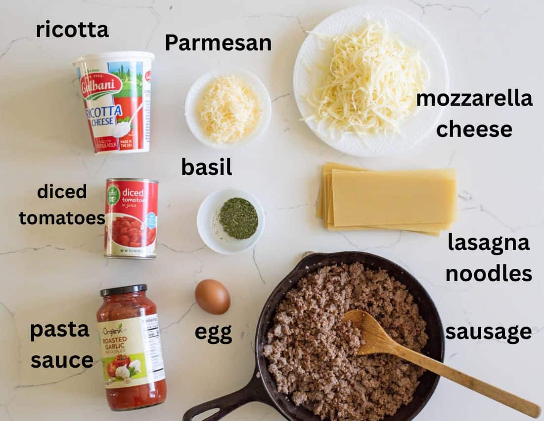 Ingredients for lasagna including ricotta, Parmesan, mozzarella cheese, diced tomatoes, basil, lasagna noodles, pasta sauce, an egg, and cooked sausage in a skillet with a wooden spoon, arranged on a white surface.
