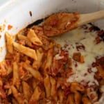 A close-up of baked penne pasta with tomato sauce and melted cheese in a white slow cooker, with a wooden spoon resting inside.