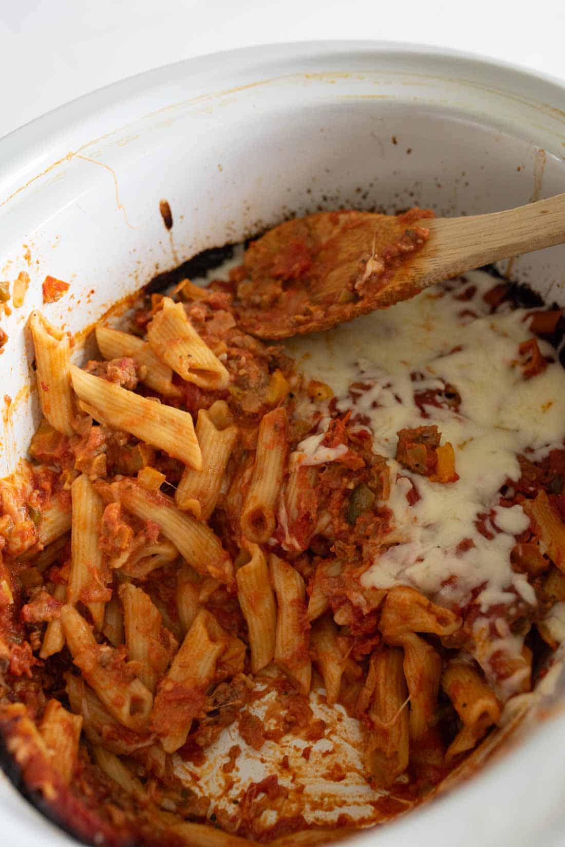 A close-up of baked penne pasta with tomato sauce and melted cheese in a white slow cooker, with a wooden spoon resting inside.