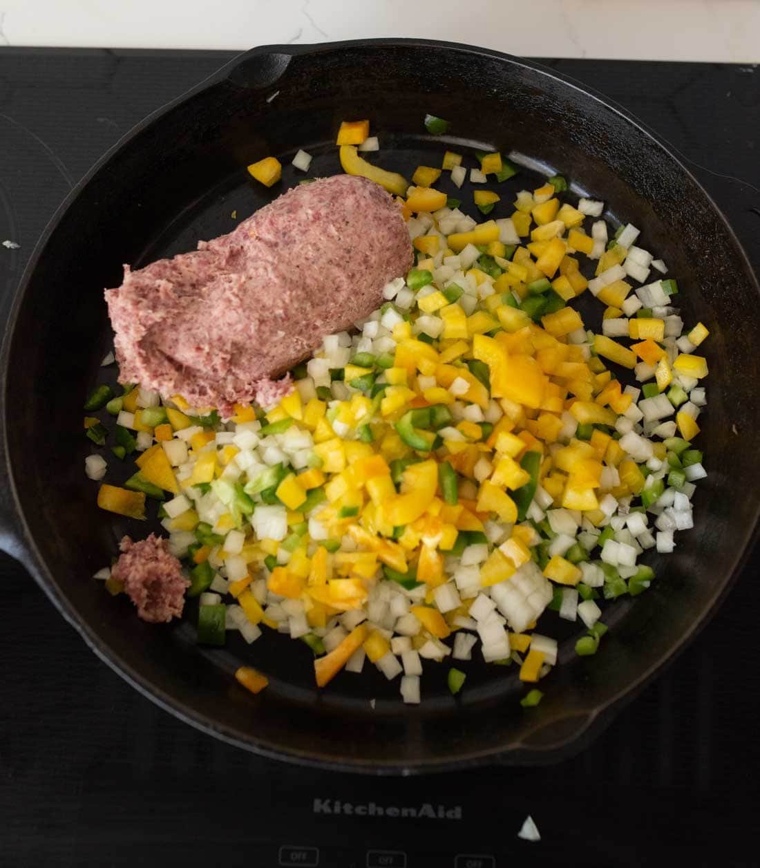 A cast iron skillet on a stovetop contains raw ground meat, chopped yellow bell pepper, green bell pepper, and diced onions, ready to be cooked.