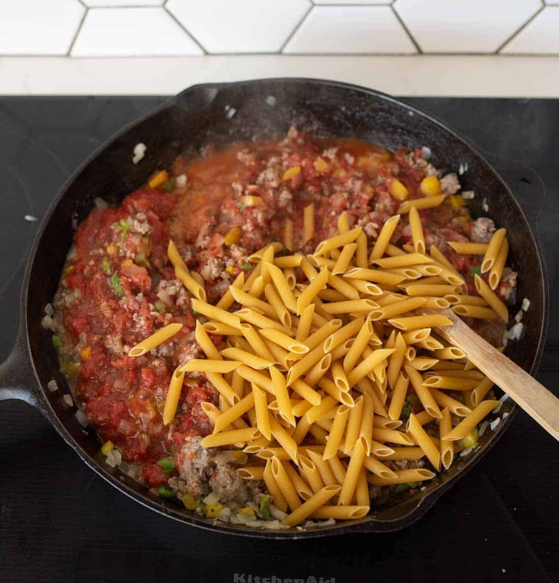 A cast iron skillet on a stovetop contains ground meat, chopped vegetables, tomato sauce, and uncooked penne pasta, being stirred with a wooden spoon. White tile backsplash is visible in the background.