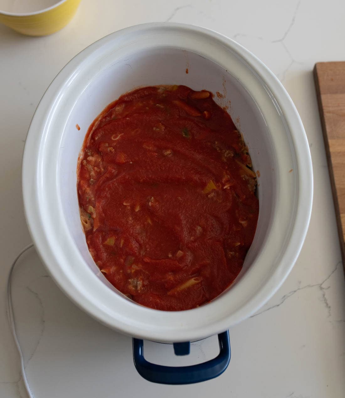 A white slow cooker filled with lasagna ingredients, topped with tomato sauce, sits on a white countertop next to a wooden cutting board.