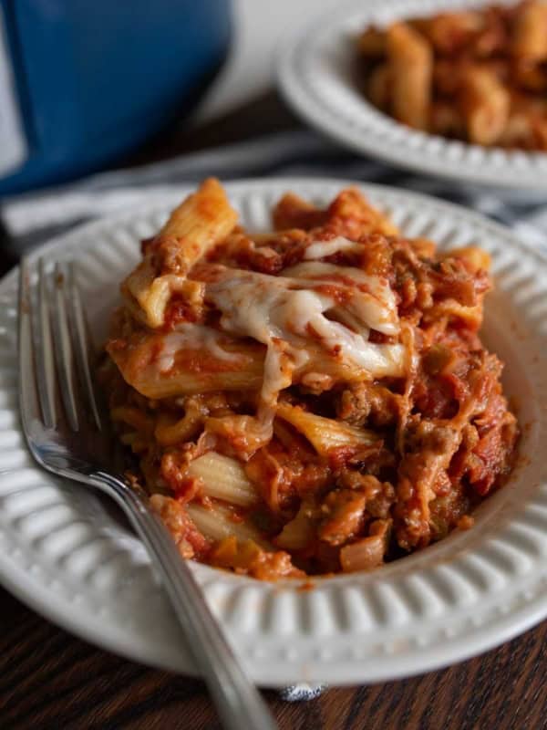 A white plate with slow cooker sausage and peppers with pasta covered in melted cheese and tomato sauce, served with a fork. Another plate of pasta and a slow cooker are visible in the background.