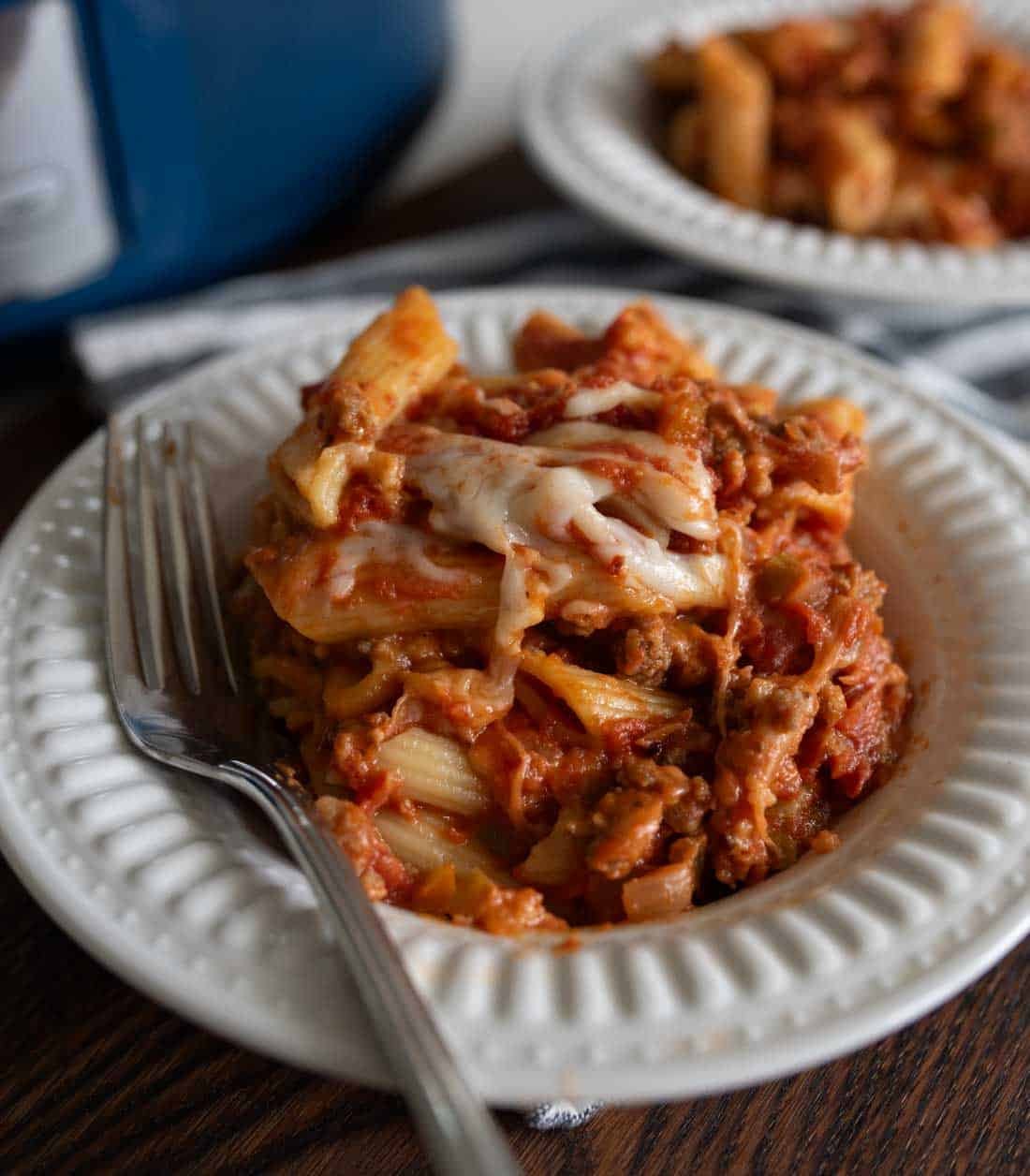 A white plate with slow cooker sausage and peppers with pasta covered in melted cheese and tomato sauce, served with a fork. Another plate of pasta and a slow cooker are visible in the background.