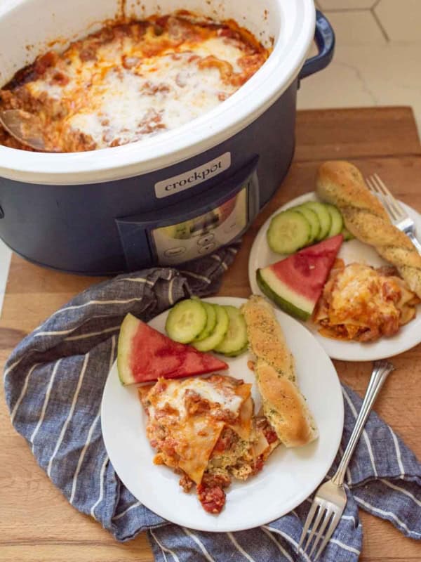 A Crockpot filled with cheesy slow cooker lasagna sits on a wooden surface. Two plates beside it hold lasagna, breadsticks, cucumber slices, and watermelon wedges. A blue striped towel lies underneath.