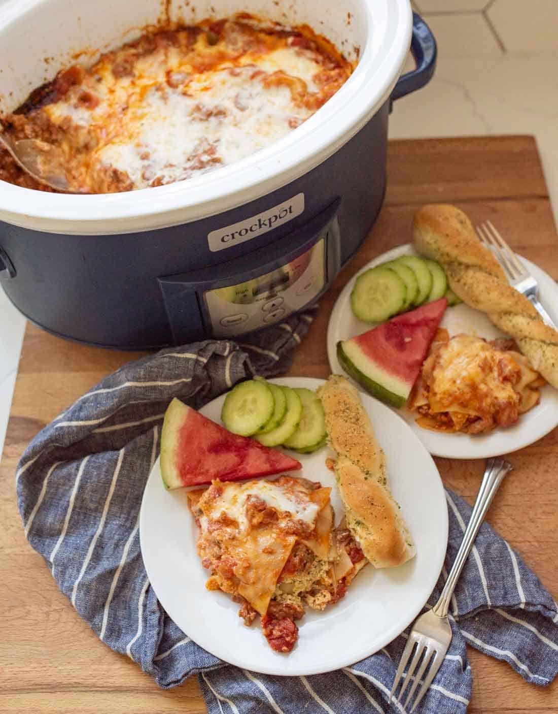 A Crockpot filled with cheesy slow cooker lasagna sits on a wooden surface. Two plates beside it hold lasagna, breadsticks, cucumber slices, and watermelon wedges. A blue striped towel lies underneath.