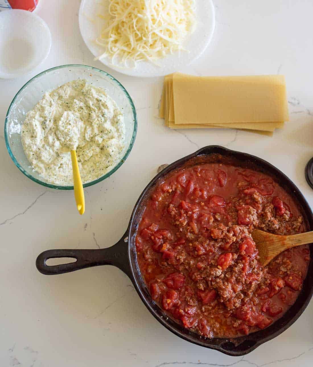 A skillet with meat and tomato sauce, a bowl of ricotta cheese mixture with a yellow spatula, dry lasagna noodles, and shredded cheese are arranged on a white countertop.