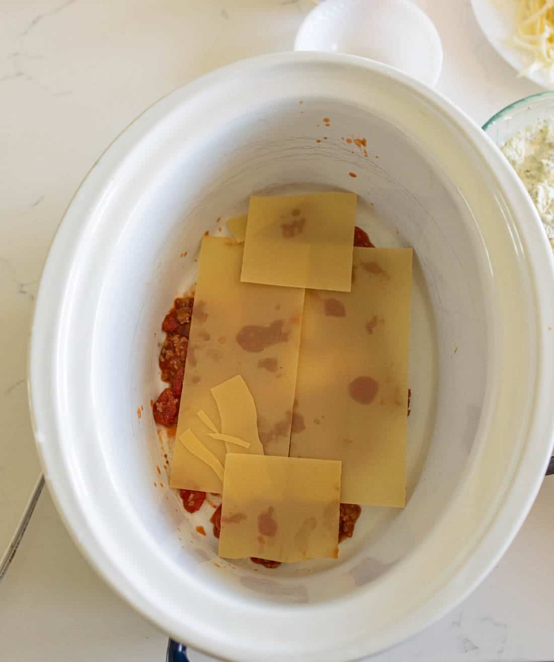 Uncooked lasagna noodles layered over a meat sauce in a white slow cooker, with broken noodle pieces filling gaps. Nearby, there are plates and bowls with additional ingredients.