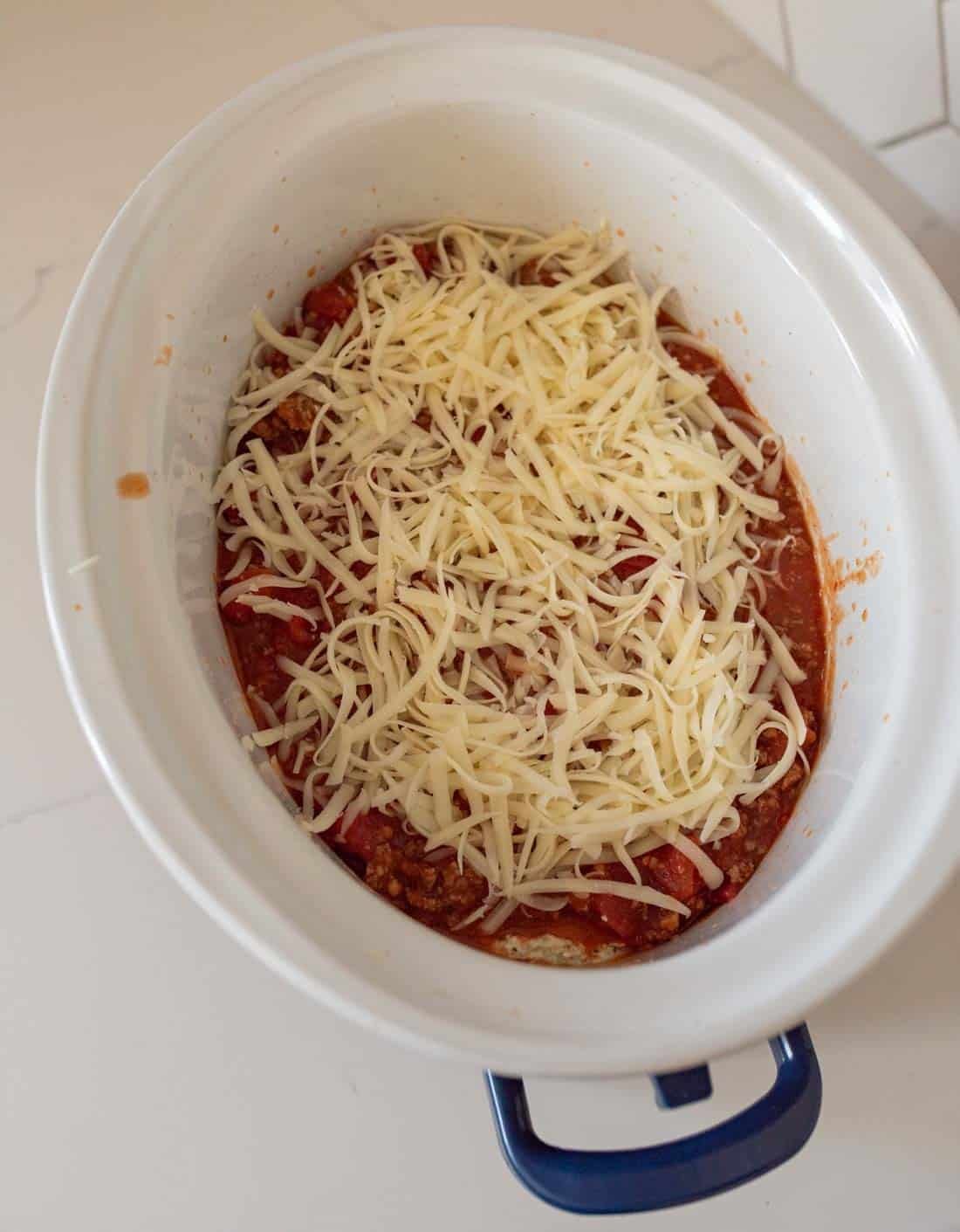 A slow cooker filled with a layer of tomato sauce, ground meat, and shredded cheese, prepared and ready to be cooked.