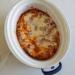 A crockpot filled with cooked lasagna, showing melted cheese and tomato sauce on top, sitting on a white countertop.