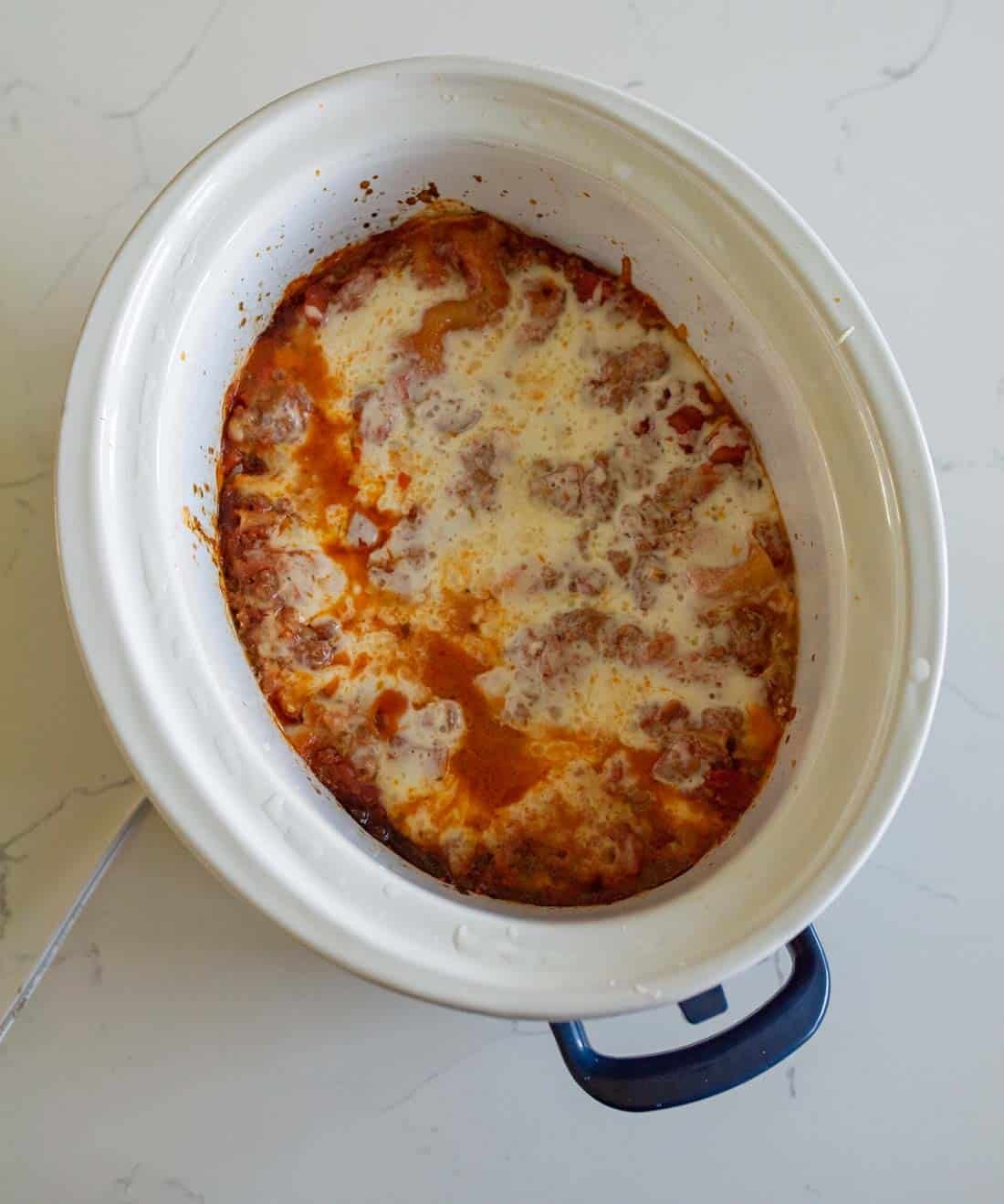 A crockpot filled with cooked lasagna, showing melted cheese and tomato sauce on top, sitting on a white countertop.