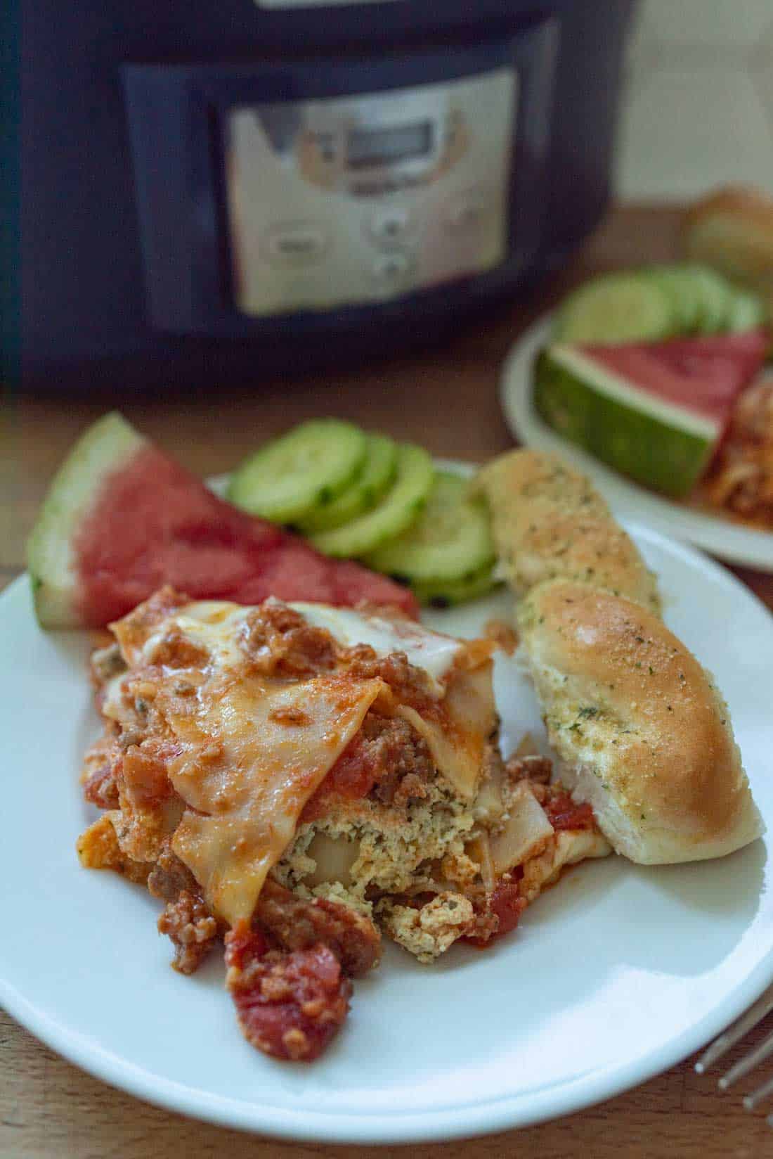 A plate with a serving of lasagna, a breadstick, sliced cucumbers, and a watermelon wedge sits in front of a slow cooker. Another plate with similar food is in the background.