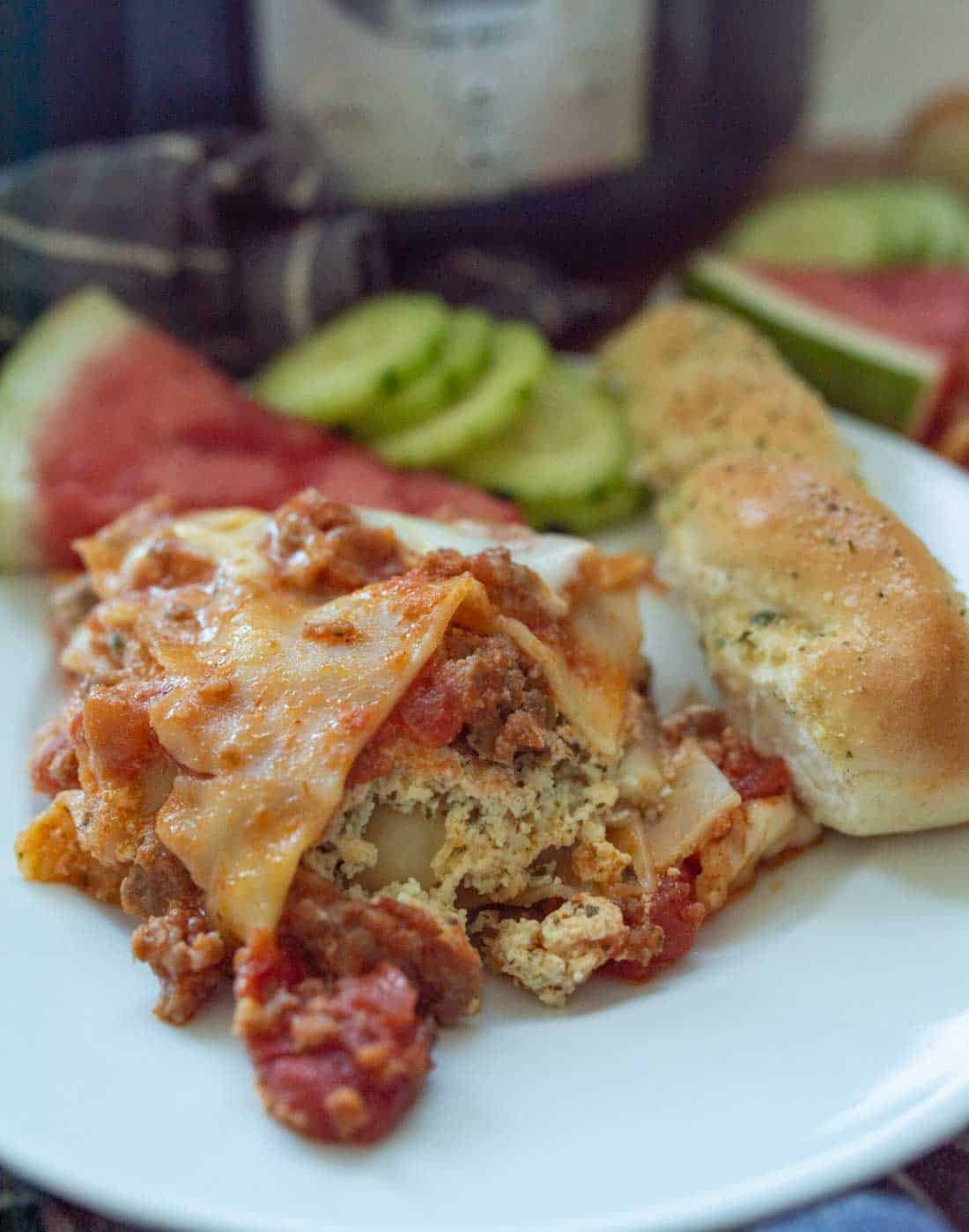 A plate with a serving of lasagna, a breadstick, sliced cucumbers, and watermelon. The lasagna has visible layers of pasta, meat, ricotta cheese, and tomato sauce. The background is slightly blurred.
