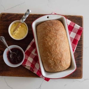 A loaf of bread in a white baking dish on a red and white checkered cloth, with bowls of butter and jam on a wooden board. A butter knife and a spoon rest in the bowls.