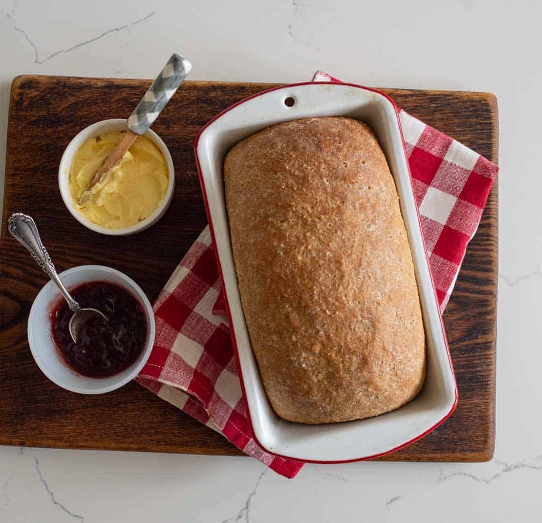 A loaf of bread in a white baking dish on a red and white checkered cloth, with bowls of butter and jam on a wooden board. A butter knife and a spoon rest in the bowls.