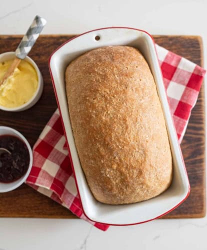 A loaf of soft whole wheat sandwich bread in a white baking dish sits on a red and white checkered cloth with bowls of butter and jam on a wooden board.