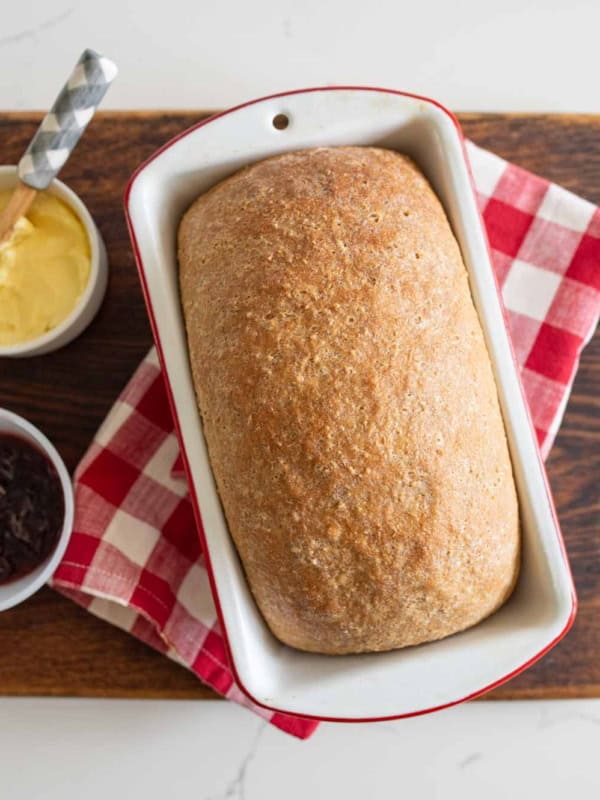 A loaf of soft whole wheat sandwich bread in a white baking dish sits on a red and white checkered cloth with bowls of butter and jam on a wooden board.