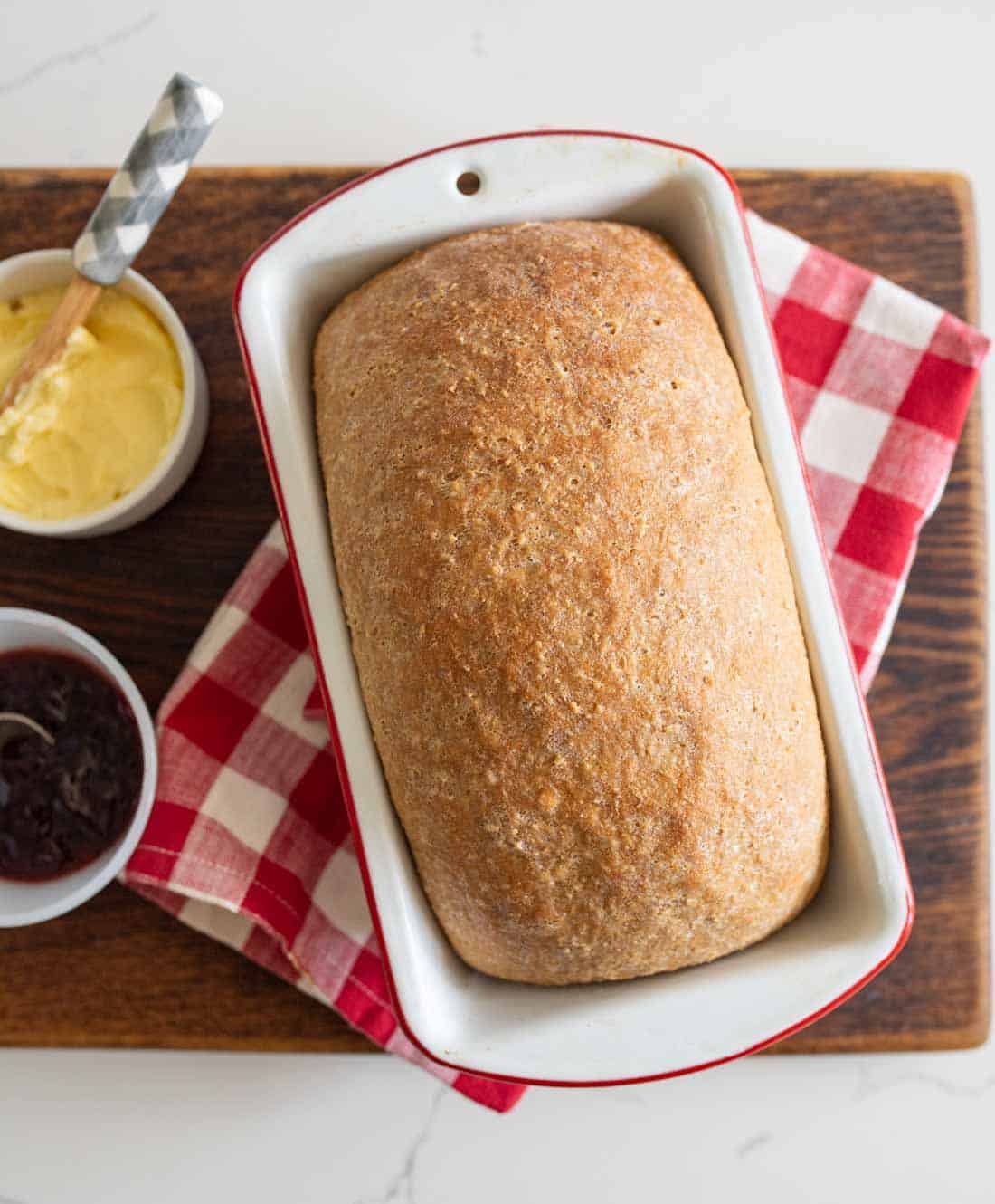 A loaf of bread sits in a white pan on a red and white checkered cloth, atop a wooden board. Next to it are small bowls of butter with a knife and dark jam.