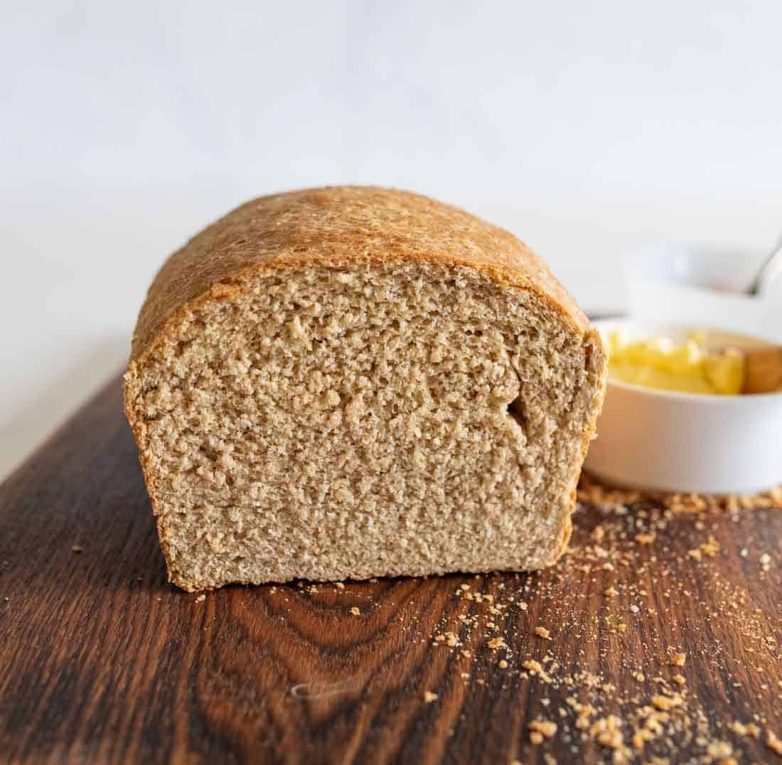 A loaf of whole wheat bread sits on a wooden surface, sliced to show its soft, dense texture. In the background, there is a white bowl with butter.