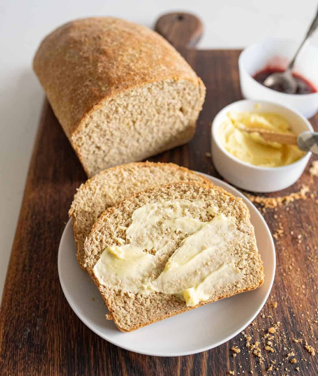 A loaf of whole wheat bread on a wooden board, with two slices on a white plate. One slice is spread with butter. Nearby are small bowls of butter and jam with a spreading knife.