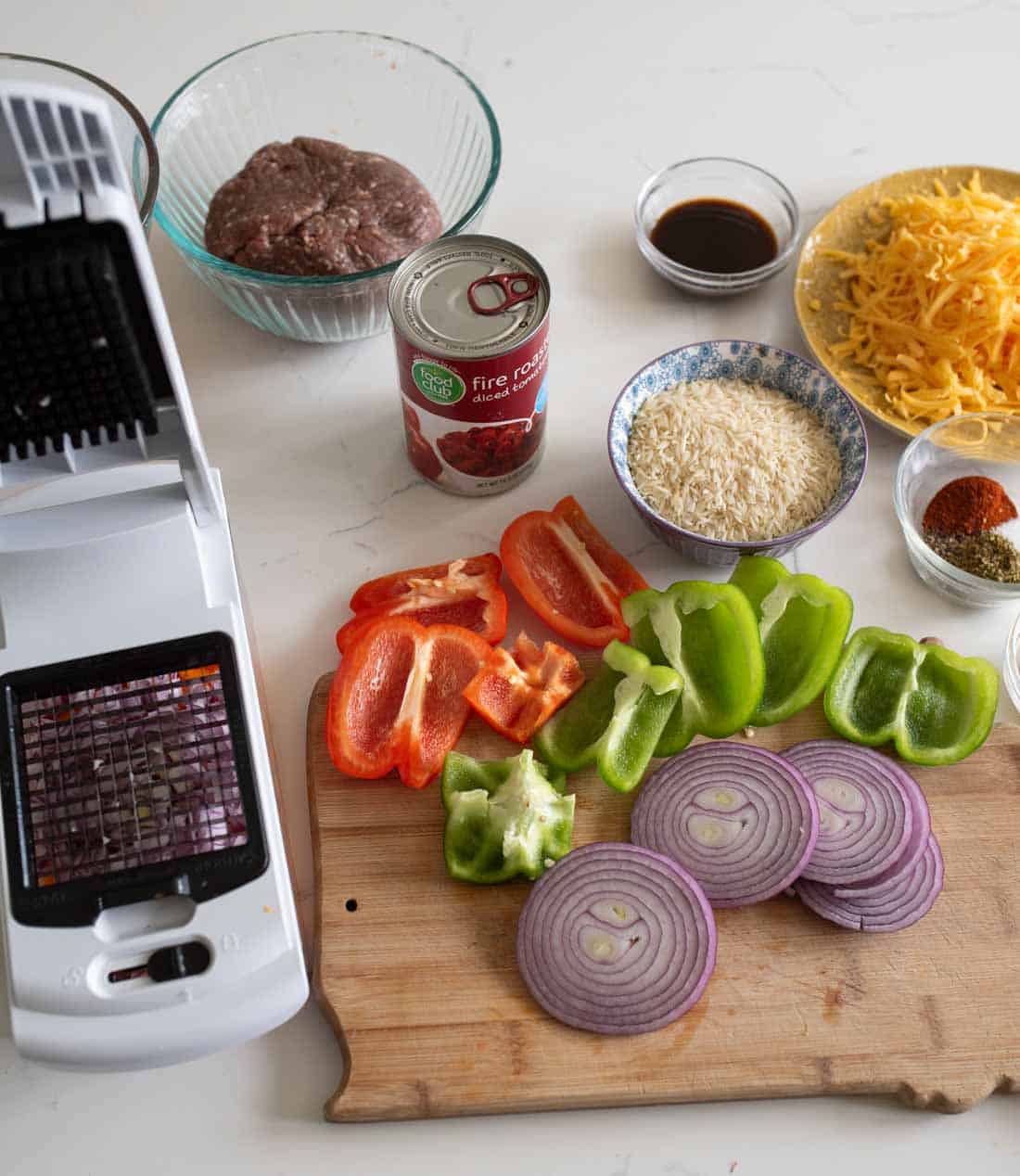 A cutting board with sliced red and green bell peppers and red onion sits next to a vegetable chopper. Nearby are bowls of ground beef, shredded cheese, rice, spices, and a can of diced tomatoes.