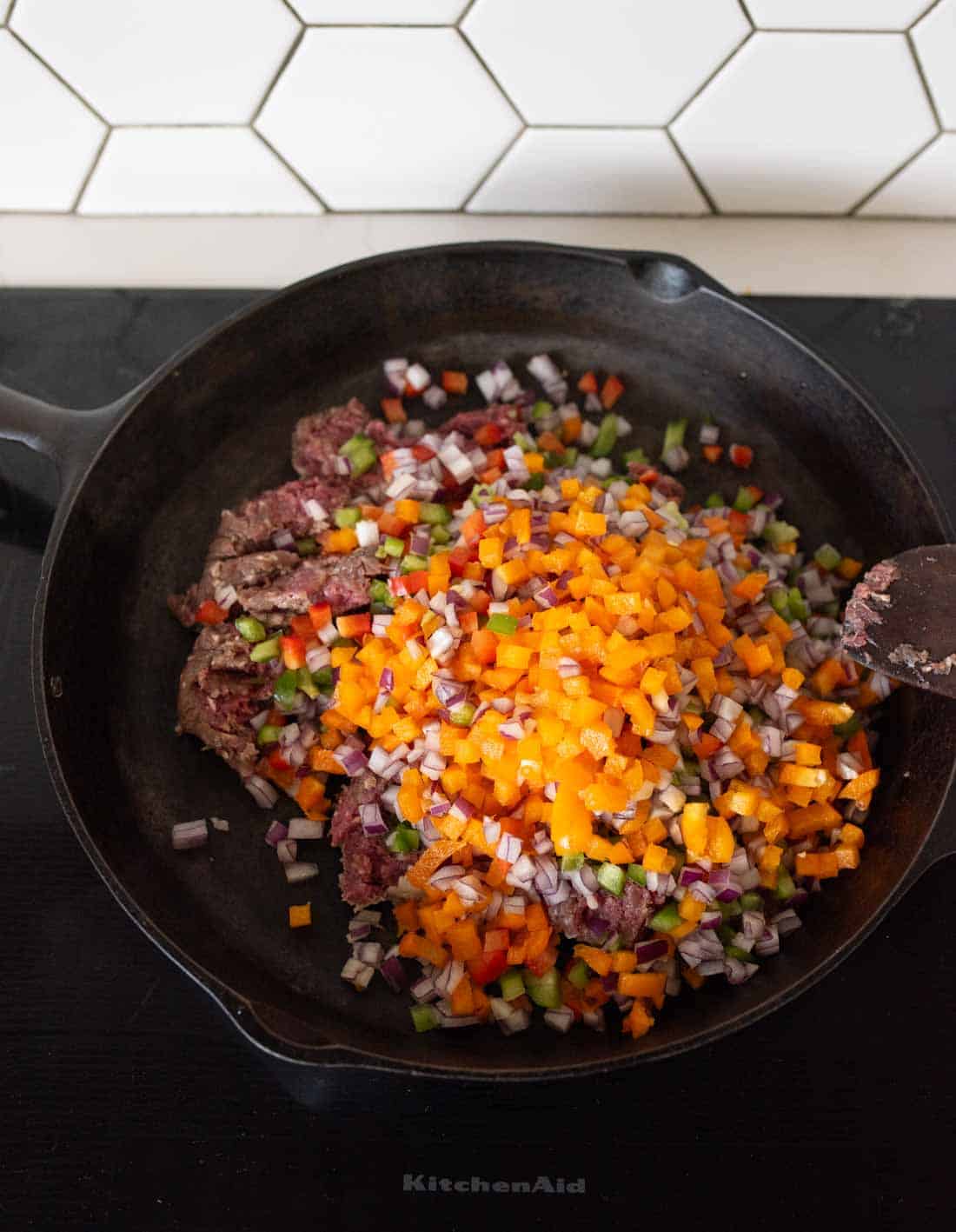 A cast iron skillet on a stovetop contains ground beef, diced red onions, green bell peppers, and chopped orange bell peppers, ready to be cooked. White hexagonal tiles are visible in the background.
