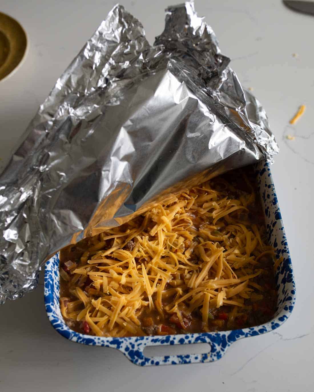 A blue and white speckled baking dish filled with shredded cheese-topped casserole is partially covered with aluminum foil on a light-colored surface.