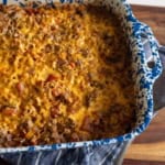 A blue and white speckled baking dish filled with a cheesy, baked casserole sits on a blue striped towel on a wooden surface. The casserole is golden brown on top with visible bits of ground meat and vegetables.