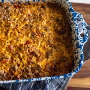 A blue and white speckled baking dish filled with a cheesy, baked casserole sits on a blue striped towel on a wooden surface. The casserole is golden brown on top with visible bits of ground meat and vegetables.