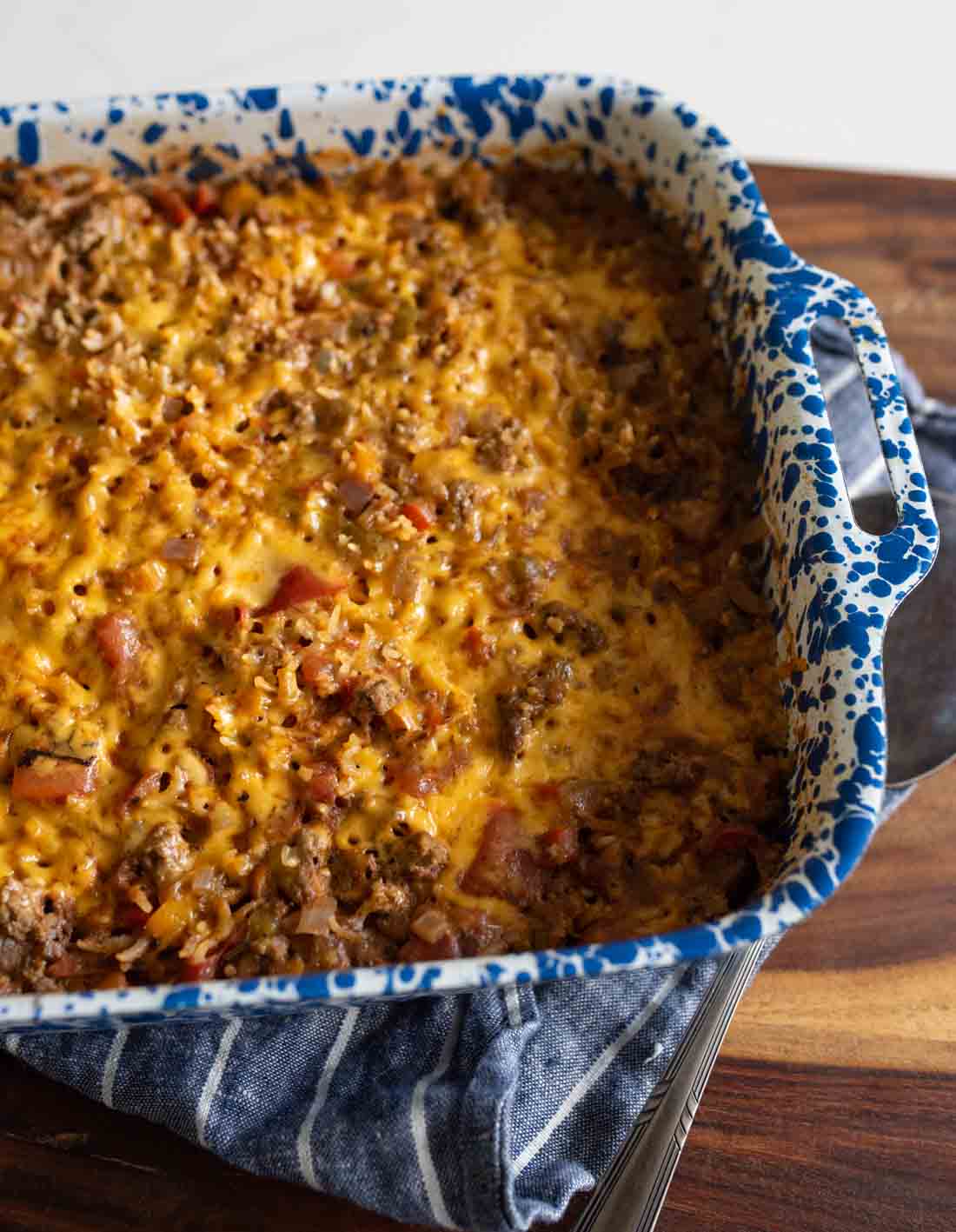 A blue and white speckled baking dish filled with a cheesy, baked casserole sits on a blue striped towel on a wooden surface. The casserole is golden brown on top with visible bits of ground meat and vegetables.