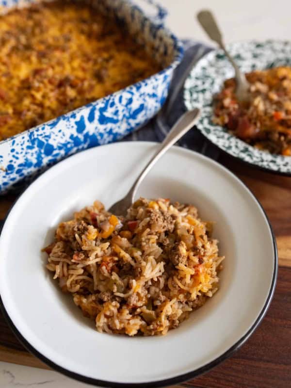 A serving of cheesy baked stuffed bell pepper casserole with ground meat and vegetables is plated in a white bowl; the casserole dish and another serving are in the background.