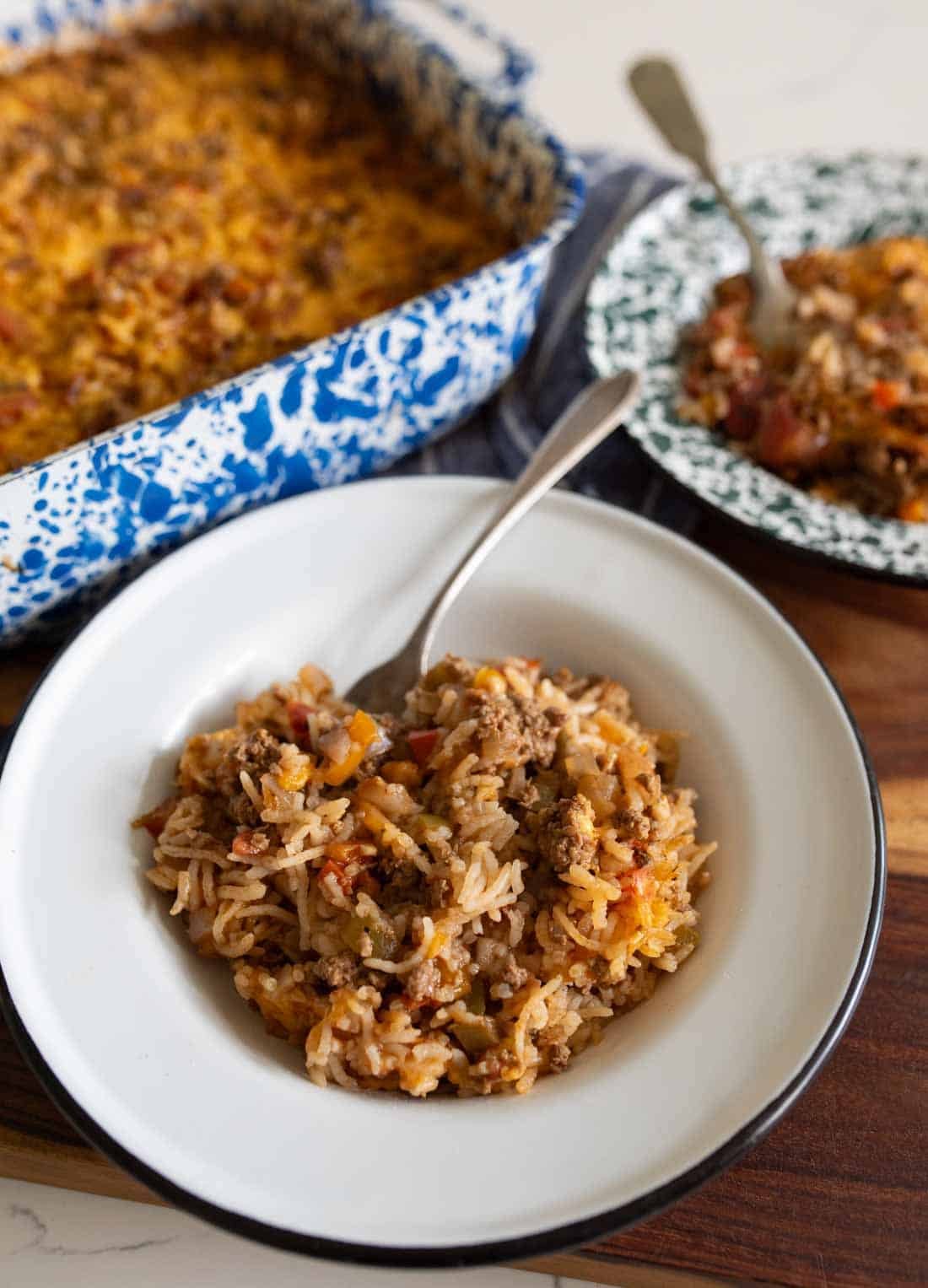 A serving of cheesy baked stuffed bell pepper casserole with ground meat and vegetables is plated in a white bowl; the casserole dish and another serving are in the background.