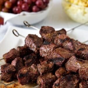 Several metal skewers with grilled beef cubes are arranged on a white plate. In the background, bowls of red grapes and yellow potato salad complement this delicious beef kebabs recipe. A website name is shown at the bottom.