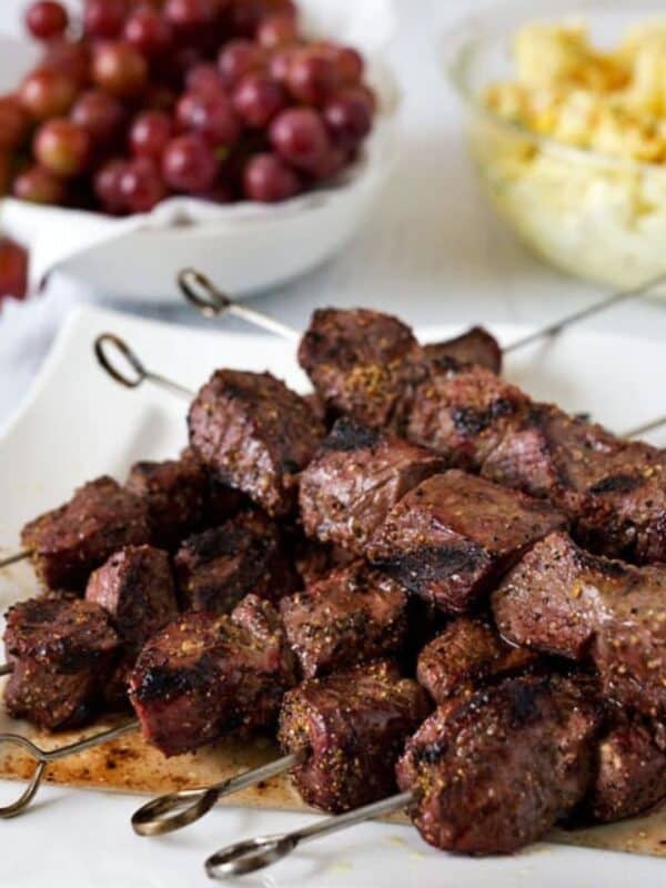 Several metal skewers with grilled beef cubes are arranged on a white plate. In the background, bowls of red grapes and yellow potato salad complement this delicious beef kebabs recipe. A website name is shown at the bottom.