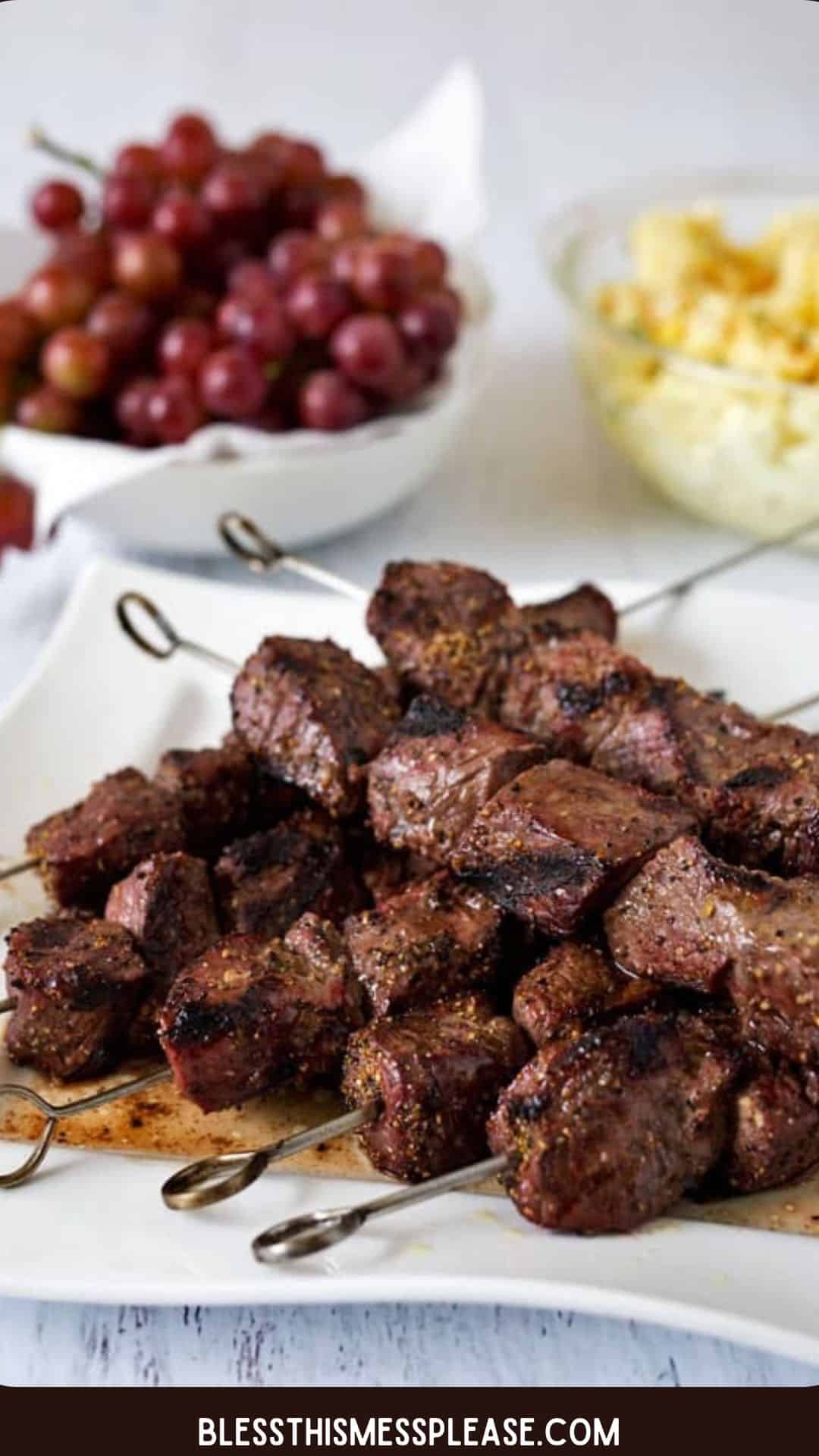 Several metal skewers with grilled beef cubes are arranged on a white plate. In the background, bowls of red grapes and yellow potato salad complement this delicious beef kebabs recipe. A website name is shown at the bottom.