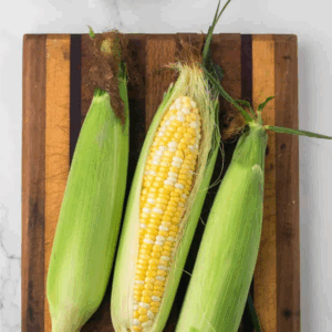 Three ears of corn, one partially husked, rest on a wooden cutting board. Nearby are salt and pepper shakers, a dish of butter, and a small bowl of oil—perfect for learning how to preserve corn at home.
